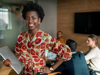 Jeune femme souriante tenant un ordinateur portable, vêtue d'une tenue colorée traditionnelle, debout devant une salle de réunion où trois collègues discutent à l’arrière-plan.